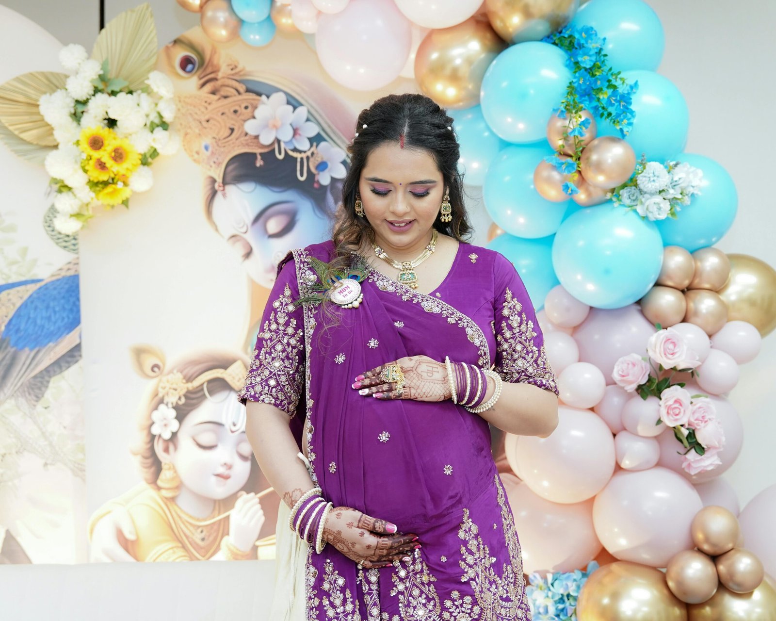 Pregnant woman at an Indian baby shower, adorned in traditional attire with festive decorations.
